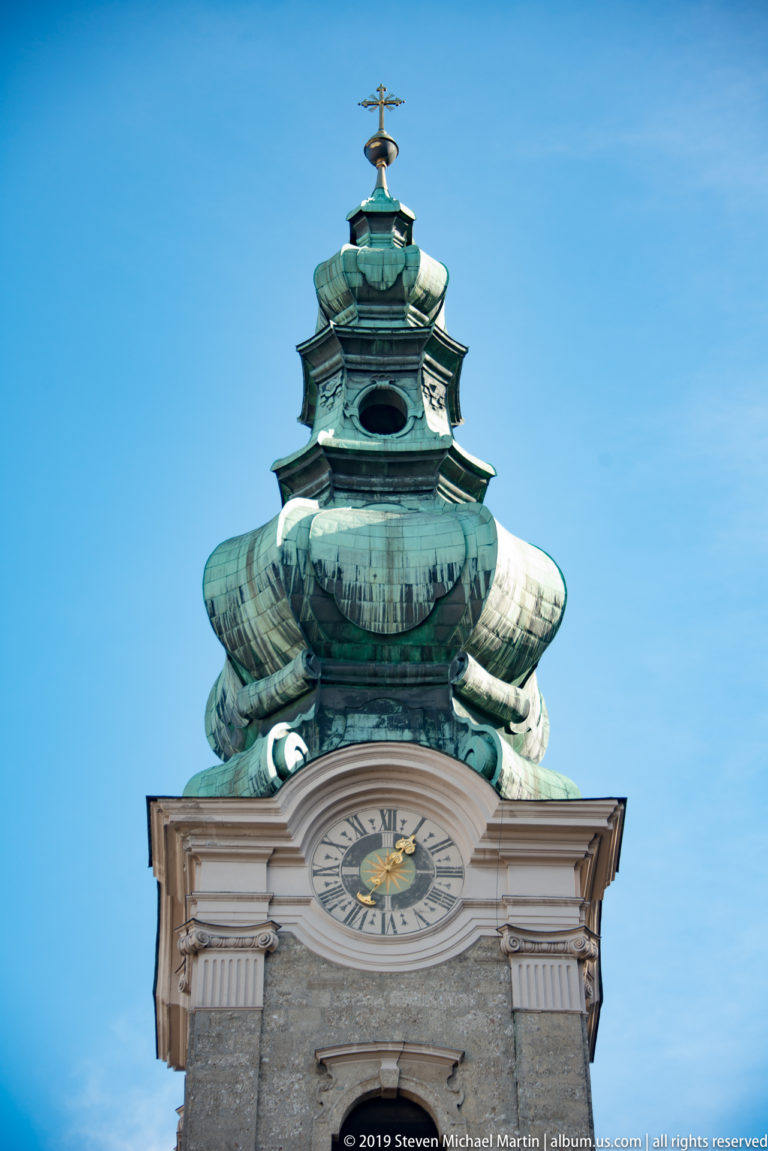 Close view of a clock face beneath the tiered green copper onion dome of St. Peter's Abbey Church tower.