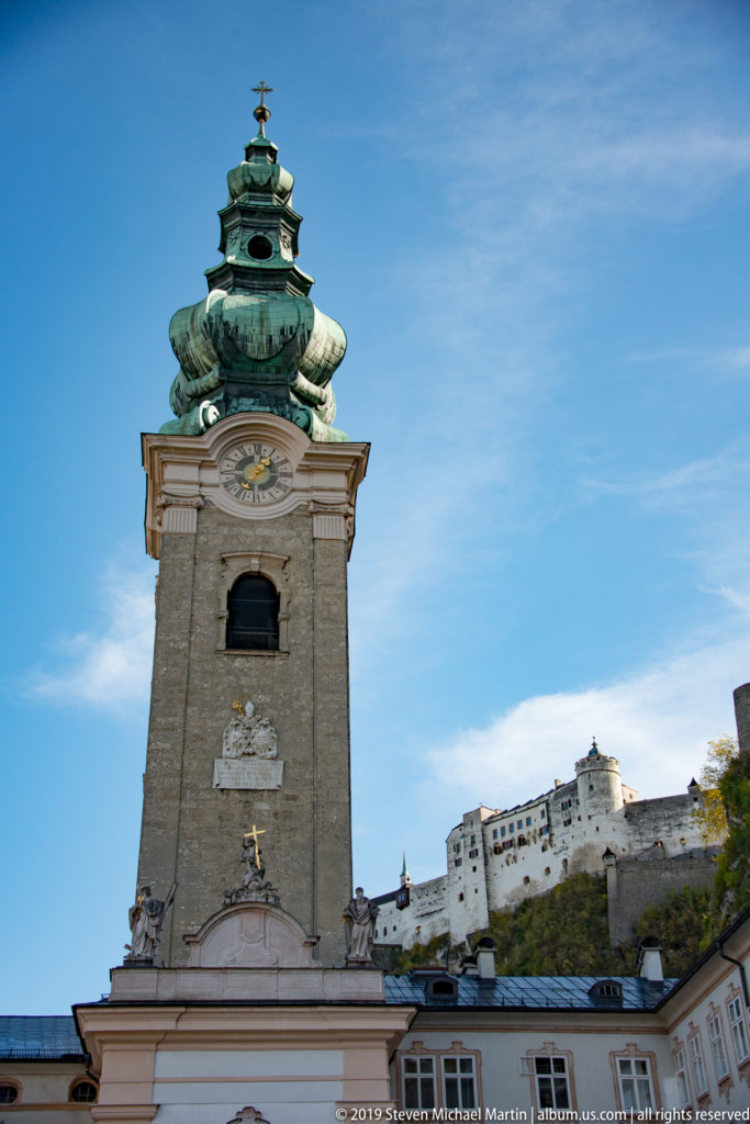 Abbey church bell tower with green onion dome in the foreground and Hohensalzburg Fortress on the ridge behind.