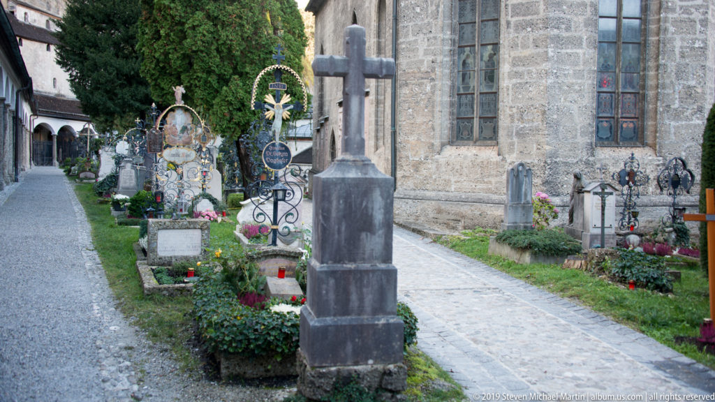 Tall stone cross beside flowered graves and iron crosses, with a church wall and large window bordering Petersfriedhof.