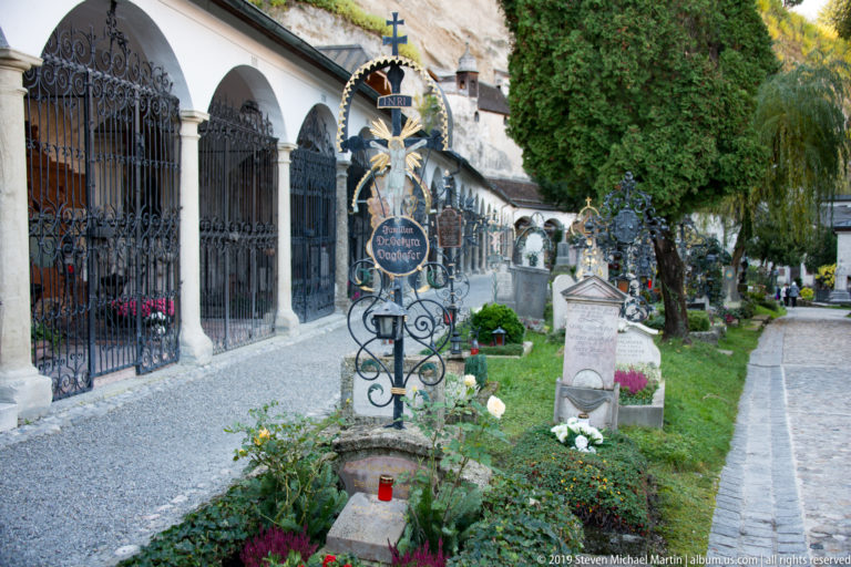 Ornate wrought-iron grave cross with crucifix and memorial plaque beside arcaded tombs and planted graves in Petersfriedhof.