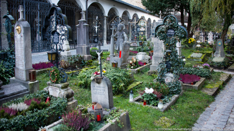 Closely spaced graves with wrought-iron crosses, stone markers, flowers, and candles in Petersfriedhof beside St. Peter’s Abbey.