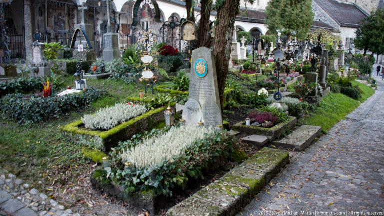 Planted graves with stone and metal memorials, flowers, greenery, candles, and decorations beside a cobbled cemetery path.