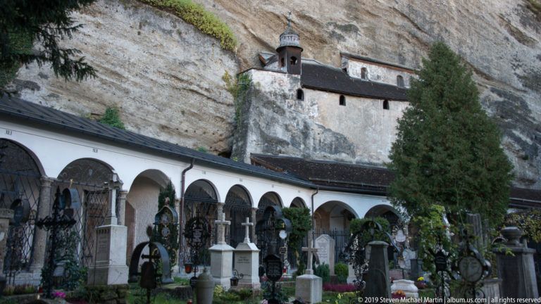 Cemetery arcades with historic grave monuments beneath a tall rock cliff and a small chapel-like structure built against the rock.