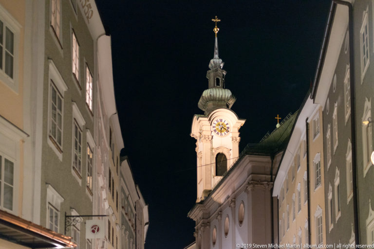Illuminated Baroque church tower with bright clock face and green onion-shaped spire between narrow Salzburg buildings at night.