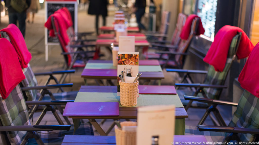 Outdoor Salzburg cafe tables with dark chairs, bright red blankets, and menu holders arranged in rows at night.