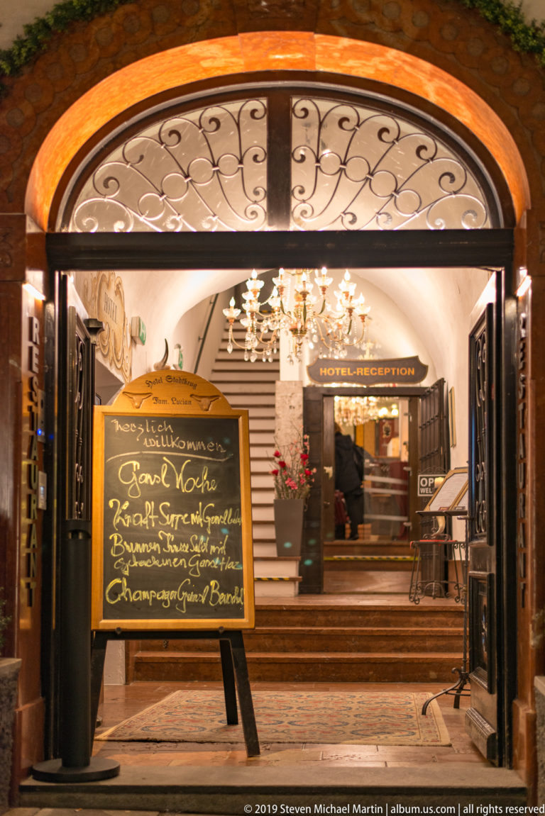 Restaurant entrance with decorative iron arch, chalkboard menu, steps, and chandelier-lit interior at night.
