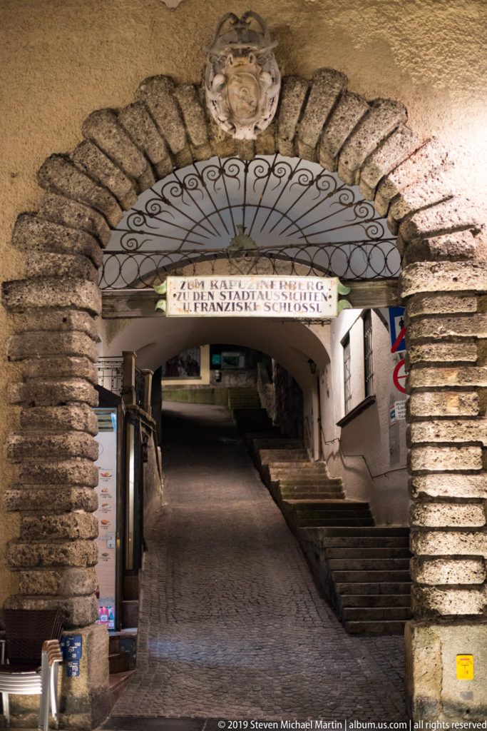 Rusticated stone archway leading into a narrow uphill Salzburg passage with steps and a decorative panel overhead.