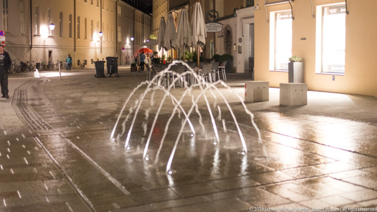 Small illuminated fountain jets arch over wet paving in a Salzburg pedestrian square with cafes and umbrellas behind.