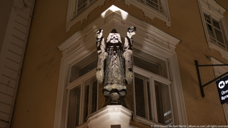 Dark vertical sculpture mounted on a building corner, lit from below against pale stucco walls and white-trimmed windows.