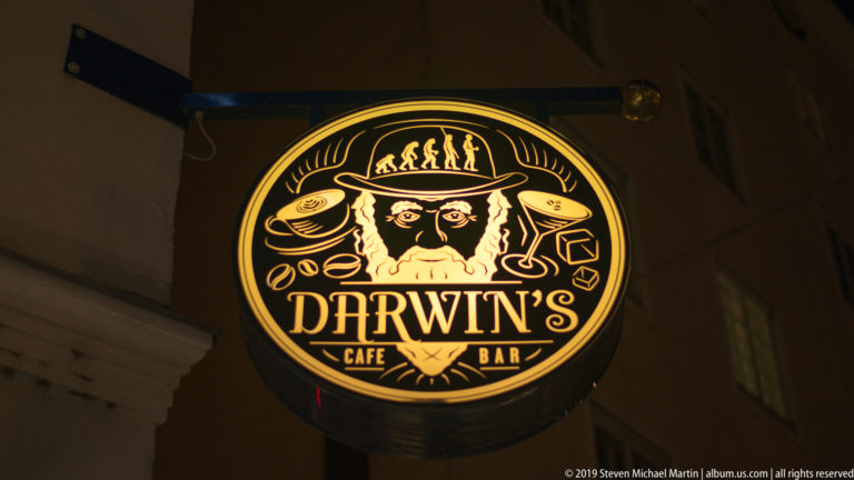 Illuminated circular Darwin's Cafe Bar sign with a stylized bearded face, evolutionary figures, and decorative symbols.