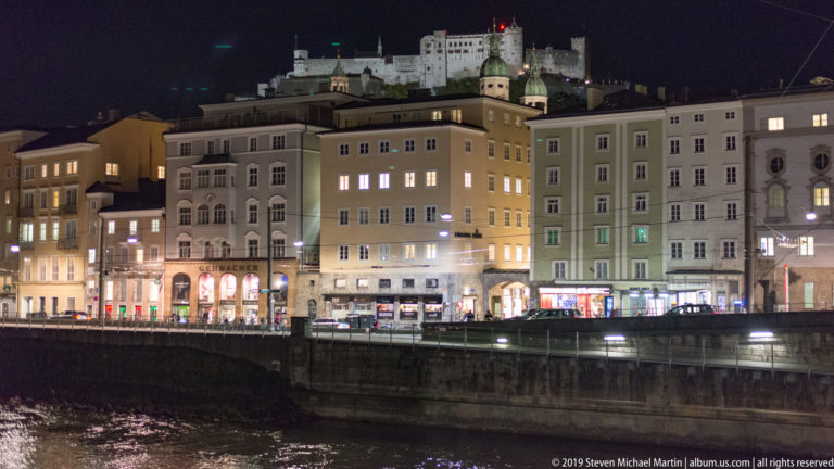Night view across the Salzach toward illuminated Salzburg buildings with Hohensalzburg Fortress high on the hill.