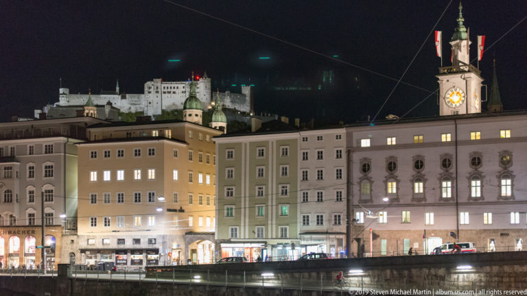 Illuminated Salzburg riverfront along the Salzach, with a clock tower and Hohensalzburg Fortress on the hill above.