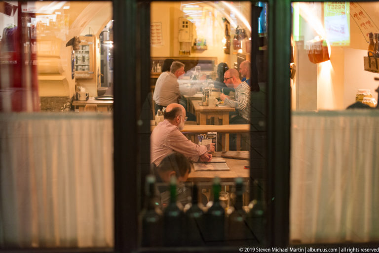 Several diners seated at wooden restaurant tables, seen through a dark-framed window with bottles along the lower edge.