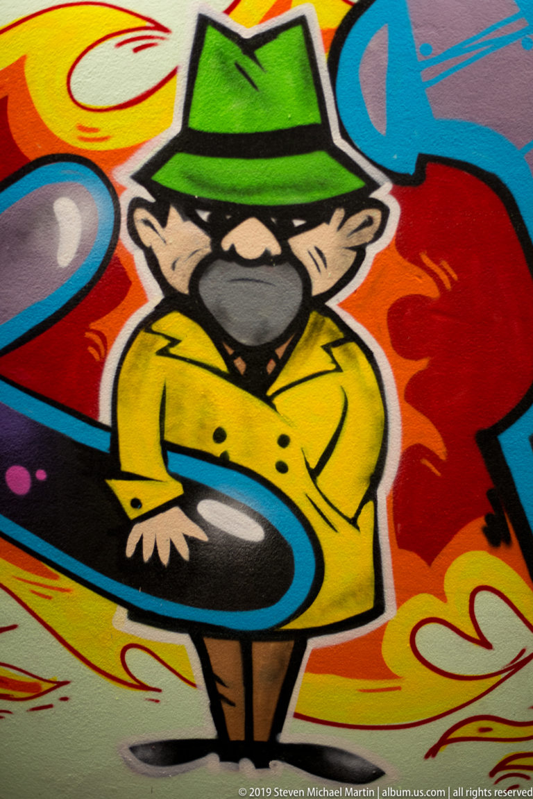 Cartoon graffiti figure in a green hat and yellow coat surrounded by bold red, blue, black, and white shapes.