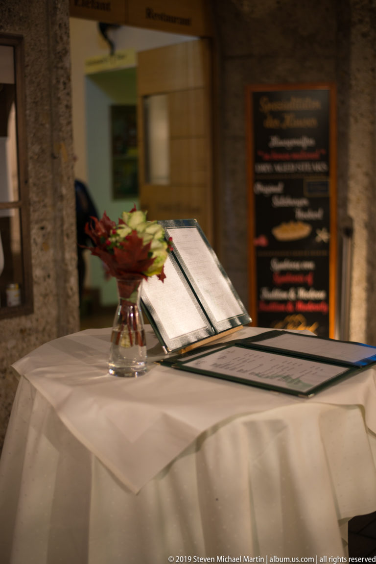 Open restaurant menu beside a small vase of red flowers on a white tablecloth, with a blurred board behind.