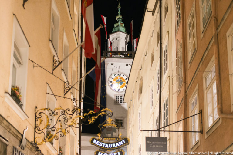 Narrow Salzburg lane with restaurant signs and Austrian flags leading toward an illuminated clock tower.