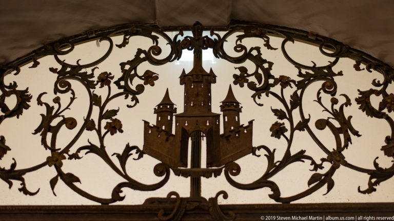 Wrought-iron hanging sign with a stylized multi-towered castle surrounded by scrolling vines and small decorative figures.