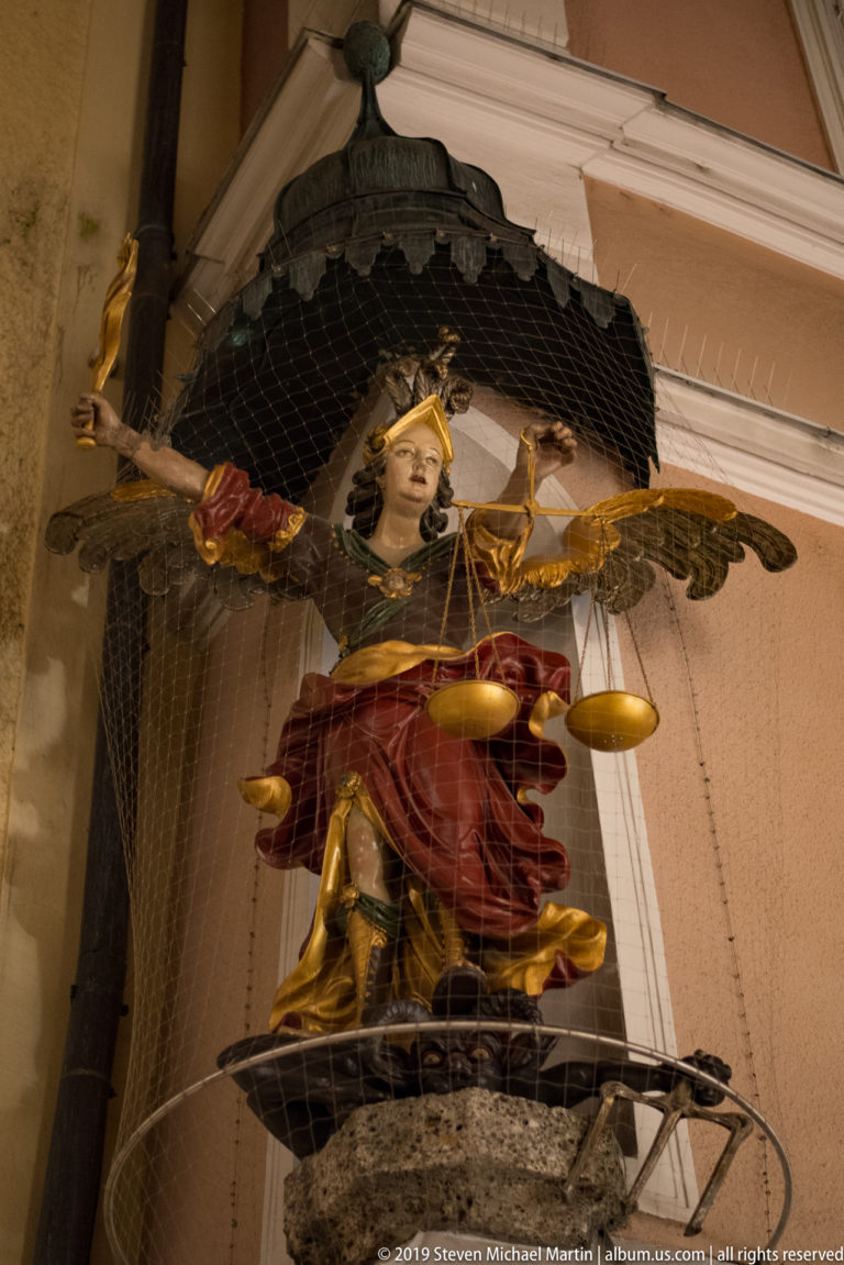 Winged armored angel figure holding balance scales beneath a canopy, with protective netting across the sculpture.