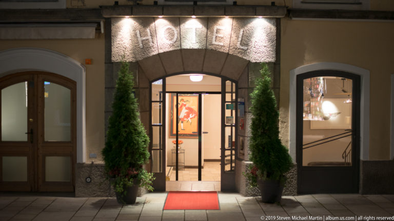 Hotel entrance at night with illuminated HOTEL letters, glass doorway, red carpet, and two potted evergreen shrubs.