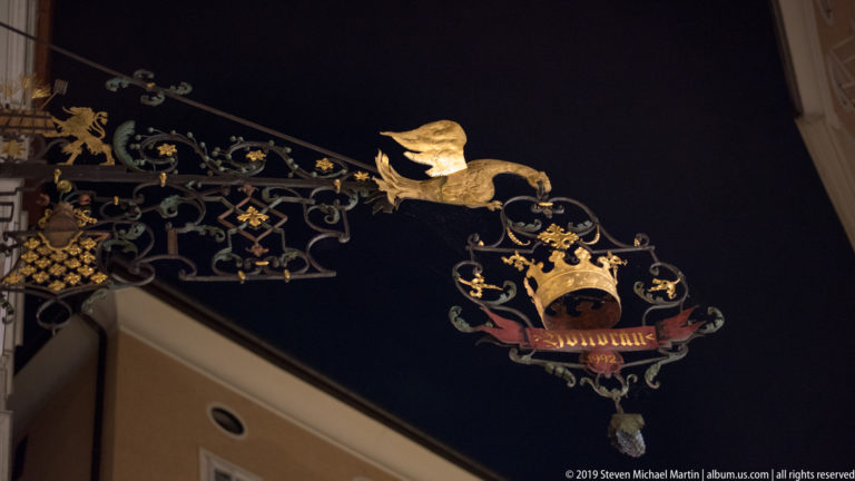 Elaborate wrought-iron hanging sign with gilded animal figure, crown, painted crest, and decorative scrollwork at night.