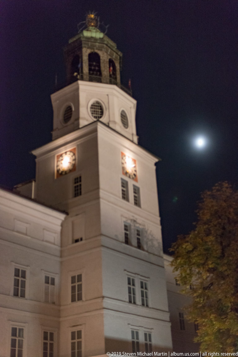 Illuminated square clock tower with two bright clock faces, green roof, and moon in the night sky.