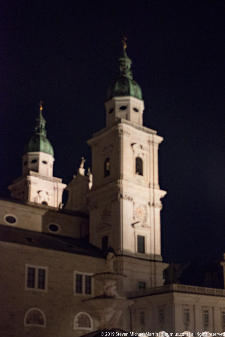Two illuminated Baroque church towers with green copper roofs rising above a lower roofline at night.