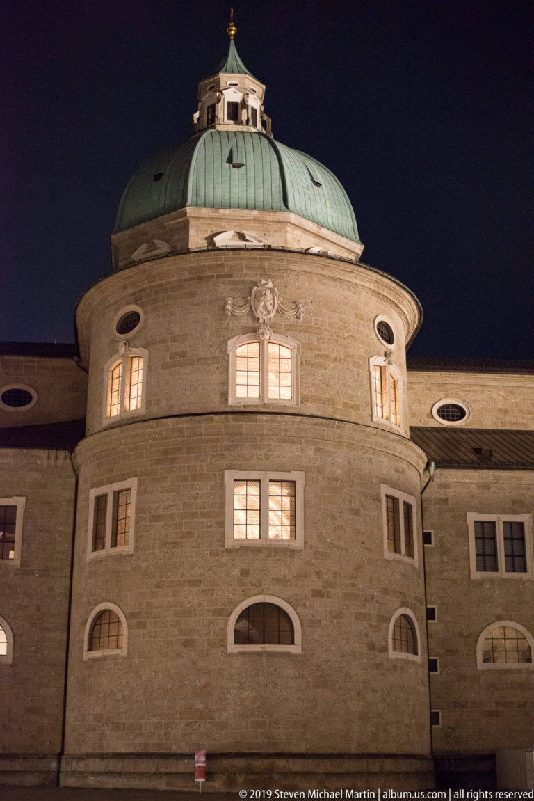 Rounded historic stone facade with lit windows beneath a broad green copper dome and small lantern at night.