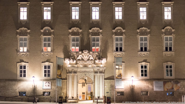 Symmetrical historic facade at night with rows of tall windows and an ornate Baroque entrance at center.