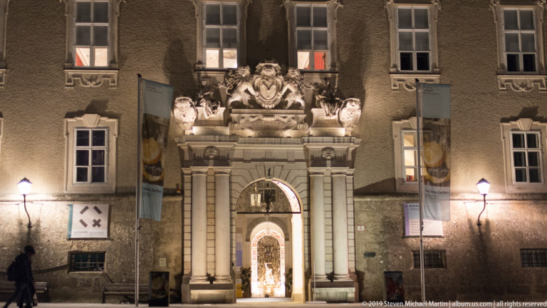 Ornate Baroque entrance with columns, sculptural crest, lanterns, banners, and a brightly lit passage beyond.
