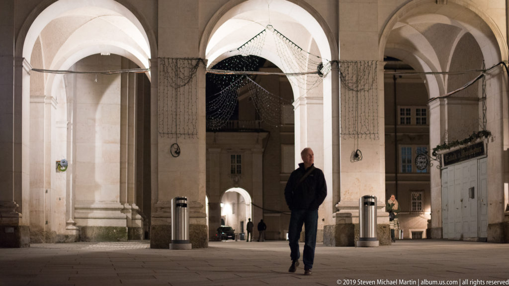 Man walking beneath tall illuminated stone arches with decorative light strings and a smaller arch beyond.