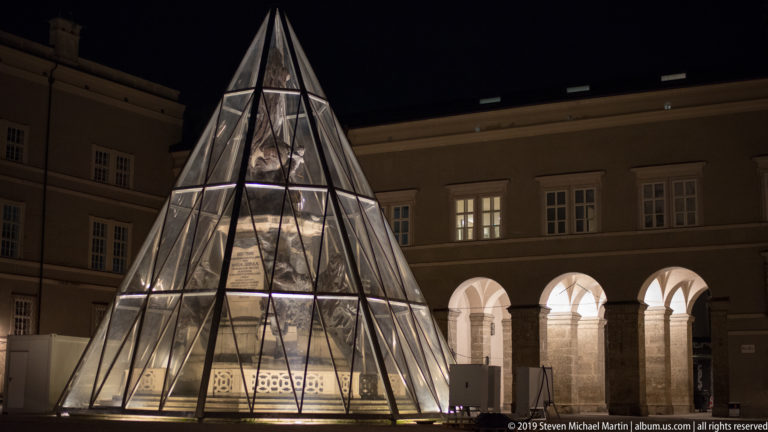 Large illuminated glass pyramid in front of a historic building with a row of bright arches at night.