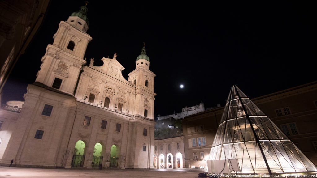 Angled nighttime view of Salzburg Cathedral with the moon, a lit glass pyramid, and Hohensalzburg Fortress beyond.