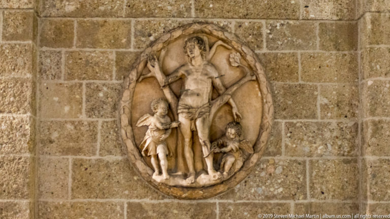 Oval stone relief of Christ on the cross with two smaller figures, mounted on a rough masonry wall.