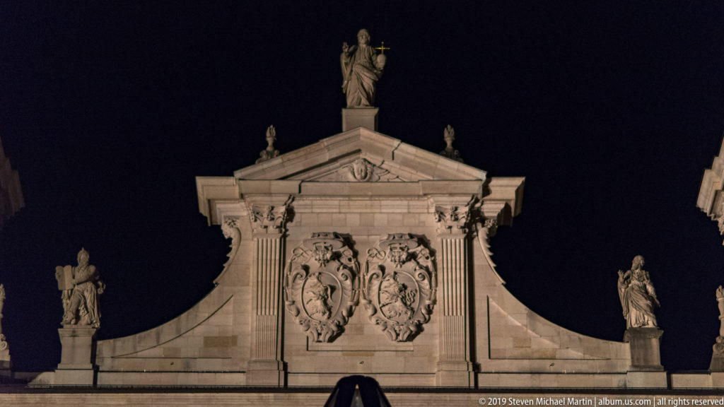 Illuminated Baroque stone pediment with central relief and statues along its curved sides and apex.