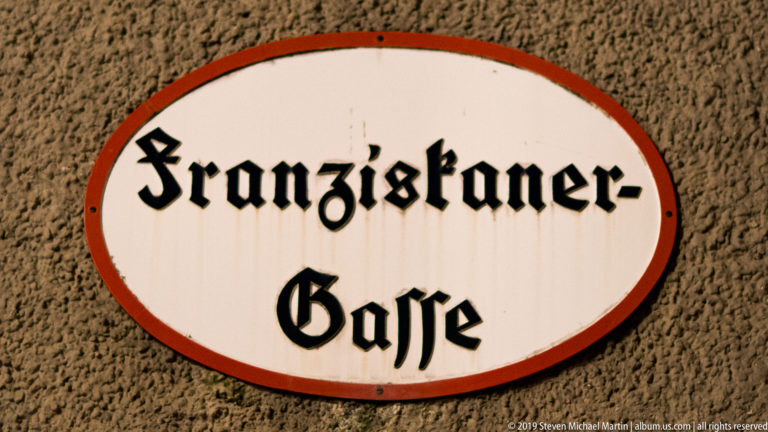 Oval white and red street plaque reading Franziskaner-Gasse in black Gothic lettering on a textured wall.