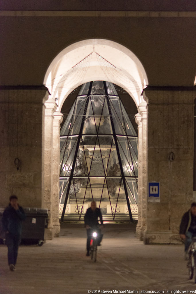 Cyclist riding beneath a large stone arch toward an illuminated glass pyramid installation at night.