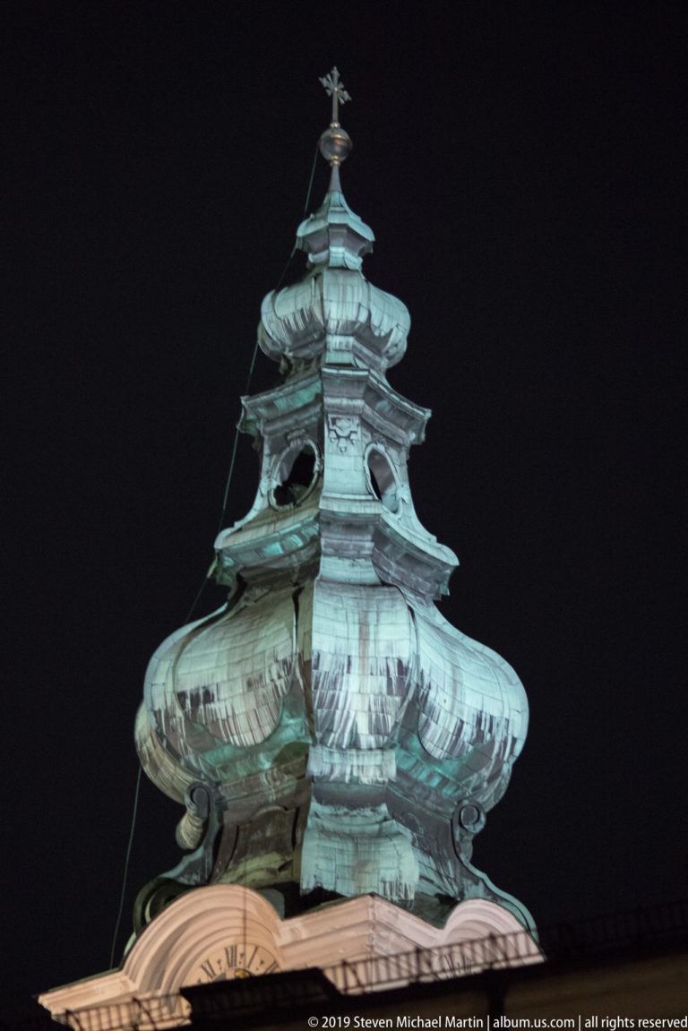 Centered close view of an ornate green copper tower roof with layered curves, openings, seams, and a small finial.