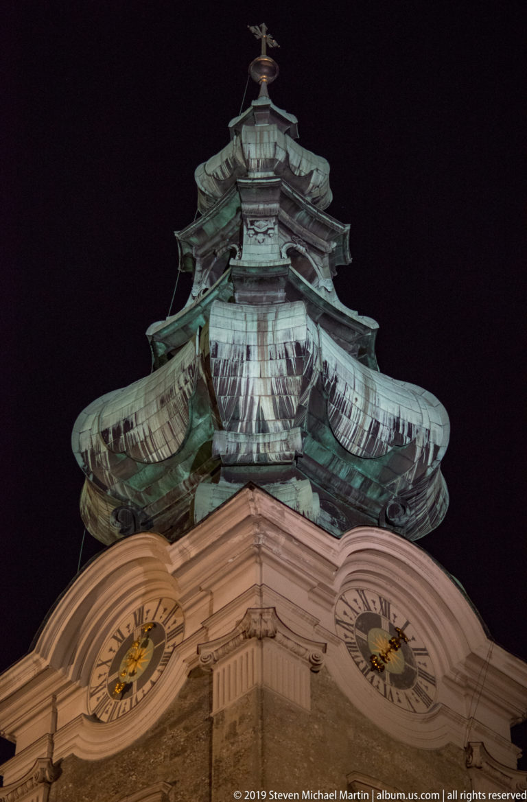 Low-angle close view of an ornate green copper tower roof above two illuminated clock faces at night.