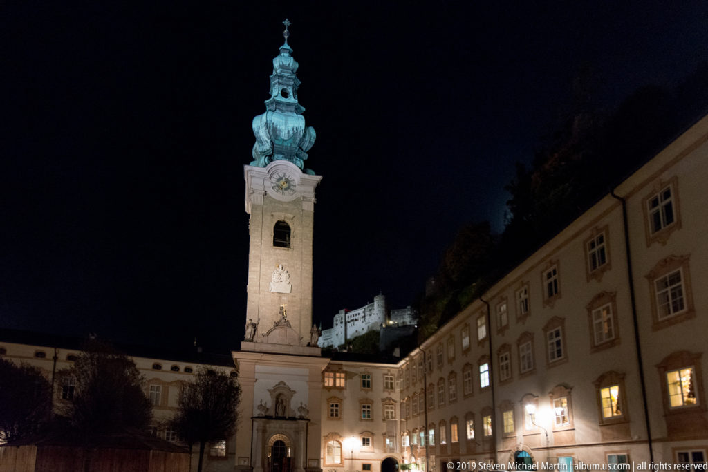 Illuminated clock tower in a Salzburg courtyard, with Hohensalzburg Fortress glowing on the hillside behind.