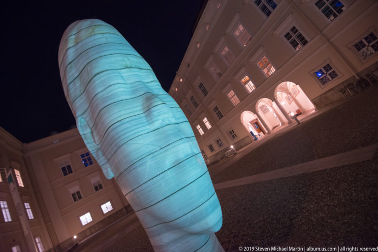 Large turquoise ribbed sculptural form rising from a historic courtyard surrounded by lit windows and arches at night.