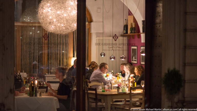 Diners seated at a candlelit restaurant table beneath a large sparkling chandelier, viewed through a window at night.