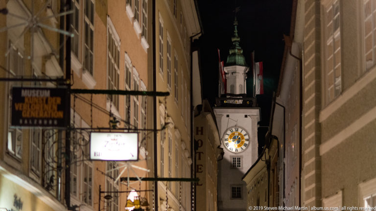 Narrow Salzburg lane framed by tall buildings and shop signs, ending at an illuminated clock tower with a bright dial.