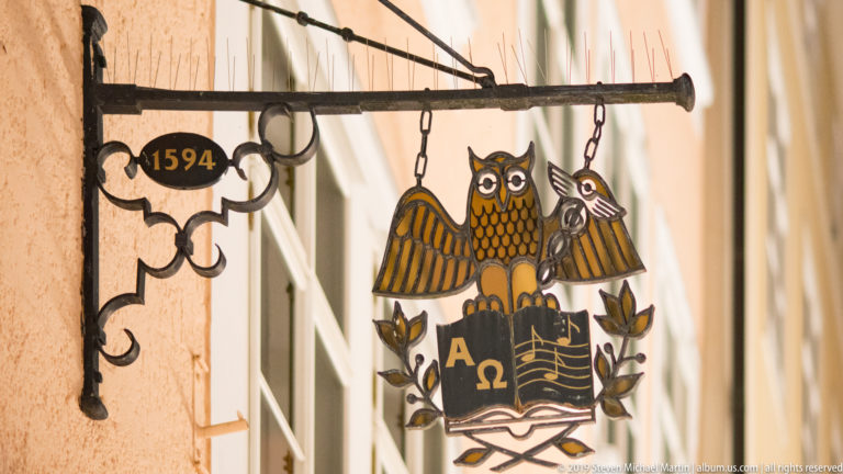 Wrought-metal hanging sign dated 1594 with an owl above an open book marked Alpha and Omega.