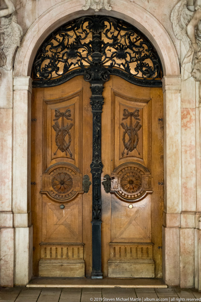 Historic double wooden doors with carved panels beneath a dense black wrought-iron arched transom.