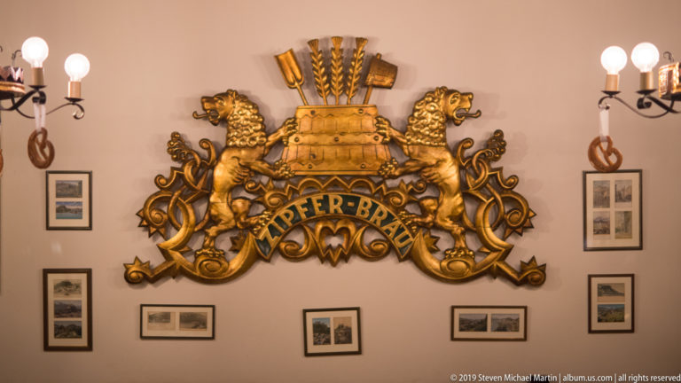 Large gilded Zipfer-Bräu wall emblem with twin lions and grain motifs above framed photographs and wall sconces.