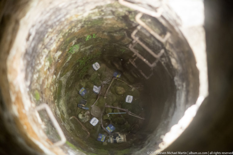 View down a deep circular stone well with a metal ladder and scattered objects visible at the bottom.