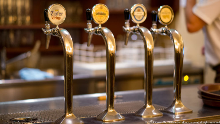 Four polished brass draft beer taps with round brand medallions lined along a bar counter.