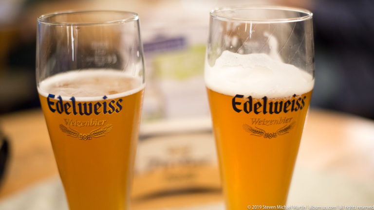 Two tall Edelweiss Weizenbier glasses with golden wheat beer and white foam on a table.