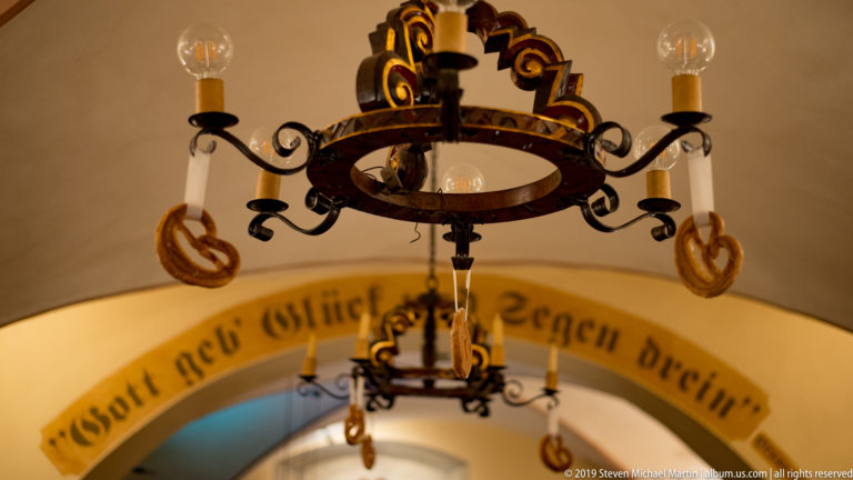 Decorative wrought-iron chandelier with curled arms and candle-style bulbs hanging before an arched German inscription.