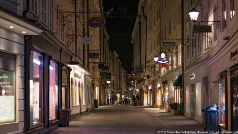 Quiet narrow Salzburg street at night with hanging shop signs, lit windows, lamps, and a few distant pedestrians.